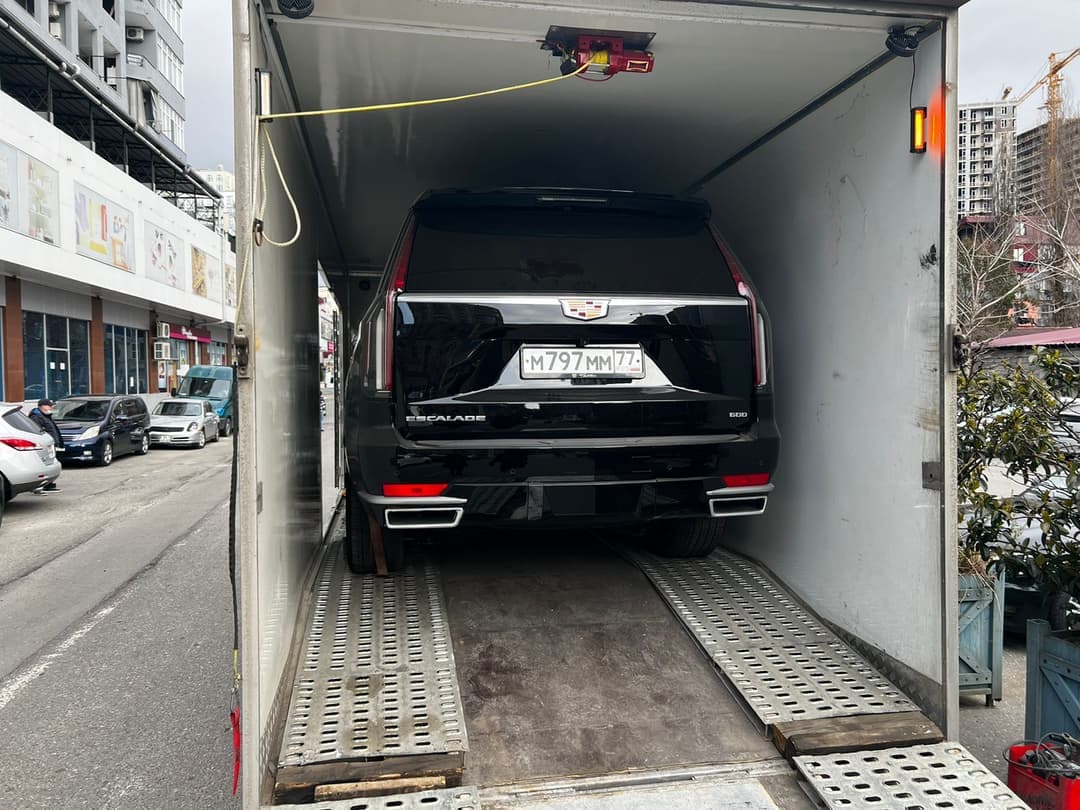 Чёрный Cadillac Escalade в закрытом кузове автовоза, вид сзади, перфорированные аппарели опущены на улицу среди жилых домов