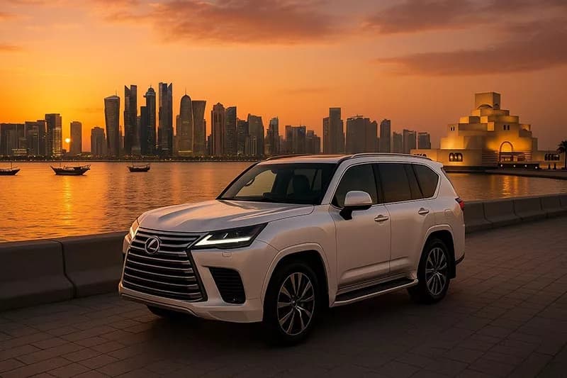 Белый Lexus LX 600 на набережной в Дохе на закате, за заливом небоскрёбы Вест-Бэй и лодки-доу, справа Музей исламского искусства