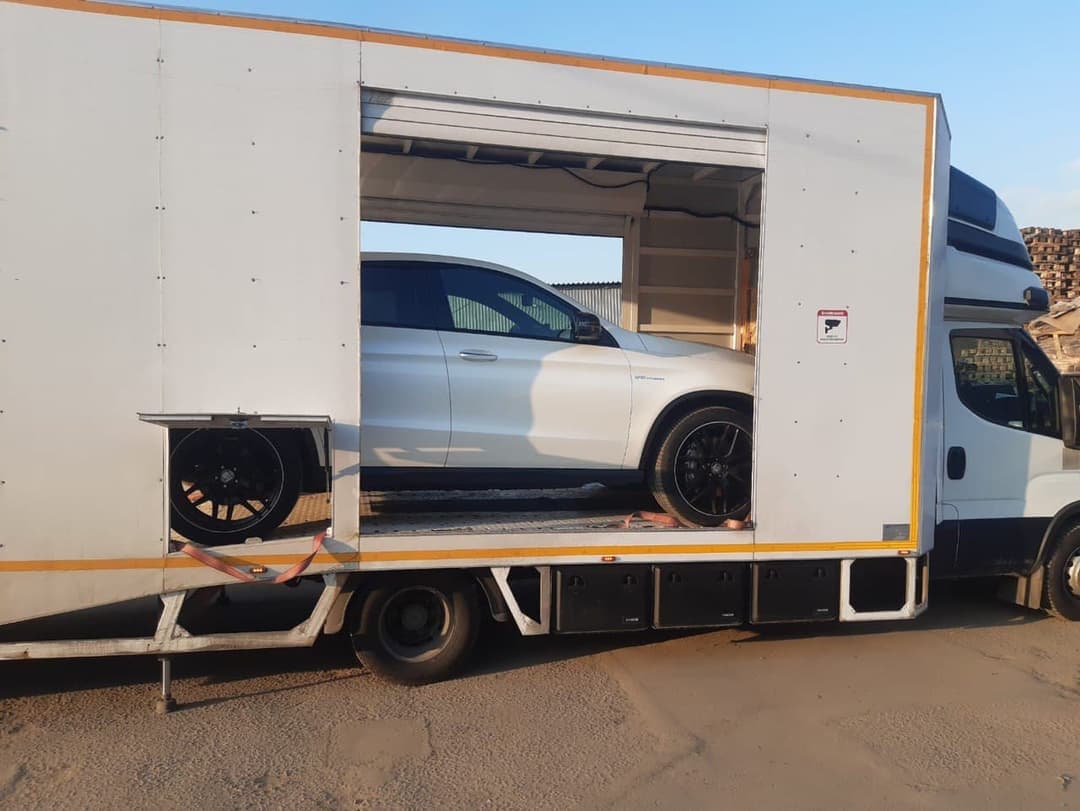 Белый Mercedes-AMG в одноместном закрытом автовозе с полностью поднятым боковым бортом, на складе позади штабеля поддонов