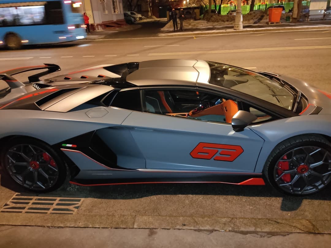 Серо-синий Lamborghini Aventador SVJ 63 Roadster с оранжевыми акцентами ночью у бордюра городской улицы